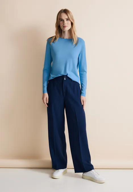 Frau mit hellblauem Pullover, dunkelblauer Hose und weißen Sneakers vor beigem Hintergrund.